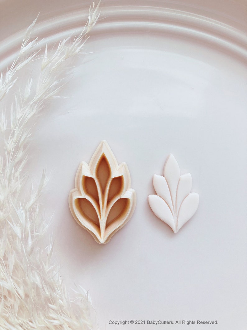 May include: Two leaf-shaped cookie cutters. One is beige and the other is white. The beige cutter has a detailed design, while the white one is a simpler version. The background is a white plate with white decorative elements.