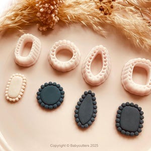 May include: A selection of white and dark teal cookie cutters in diverse shapes, such as ovals, circles, and rectangles, with decorative beaded edges. The cutters are displayed on a light pink plate, with dried ornamental grass in the background.
