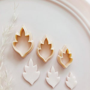 May include: A collection of leaf-shaped cookie cutters and fondant cutouts in different sizes. The cutters are beige, and the cutouts are white, displayed on a pale pink plate. The image has a soft, pastel aesthetic, ideal for baking and crafting projects.