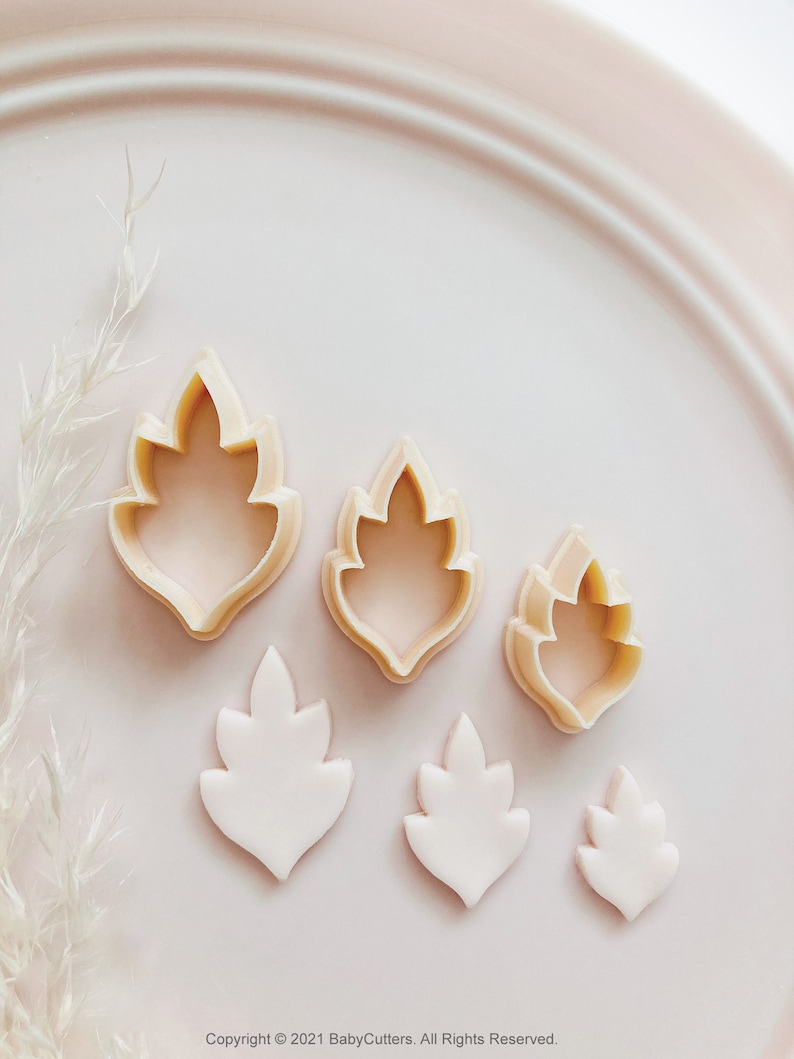 May include: A collection of leaf-shaped cookie cutters and fondant cutouts in different sizes. The cutters are beige, and the cutouts are white, displayed on a pale pink plate. The image has a soft, pastel aesthetic, ideal for baking and crafting projects.