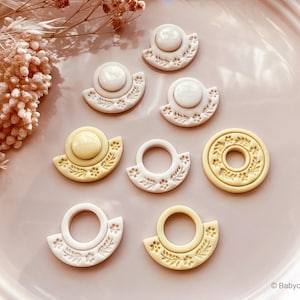 May include: A collection of decorative cookie cutters in various shapes and colors. The cutters include a hat, a ring, and a circular design, all with floral patterns. The colors are white, cream, and yellow. The items are arranged on a pink surface.