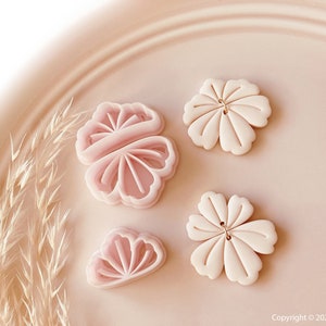 Puede incluir: Cuatro cortadores de galletas rosas con forma de flor sobre un plato rosa claro. Dos cortadores son flores abiertas y dos están cerradas. Los cortadores tienen diseños detallados de pétalos.