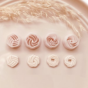 May include: Eight light pink cookie cutters in floral shapes. The cutters are arranged in two rows of four, featuring spiral and woven designs. The background is a matching light pink, with a few sprigs of dried grass. Copyright 2023 Babycutters.