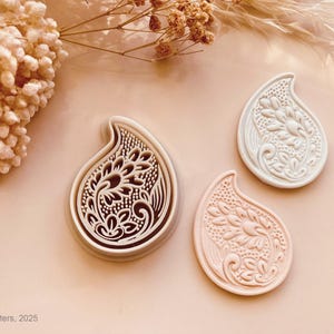 May include: Three paisley-shaped cookie cutters with intricate floral designs. One is a cookie cutter, and two are cookies. The cookie cutter is white with a brown outline. The cookies are white and light pink. The background is a light peach color.