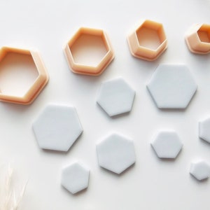 May include: A selection of hexagon-shaped cookie cutters and cut-out shapes. The cutters are a light peach colour, and the cut-out shapes are white. The sizes vary, with the largest cutter and shape at the top and decreasing in size.
