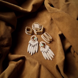 Peut inclure: Vue aérienne de plusieurs emporte-pièces crème et de deux paires de boucles d'oreilles à motif de plumes. Les boucles d'oreilles ont un accent de pierre turquoise. Les articles sont disposés sur un tissu marron texturé.