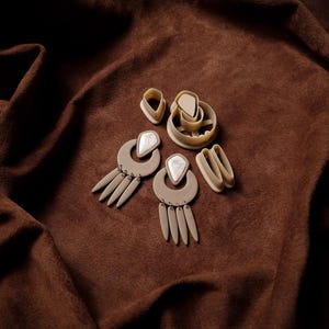 Peut inclure: Des boucles d'oreilles en argile polymère beige et des outils de fabrication de bijoux sont disposés sur un tissu marron. Les boucles d'oreilles ont une forme de croissant avec des éléments pendants et une incrustation de pierre blanche.