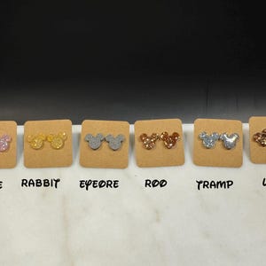 May include: Six pairs of glitter mouse ear stud earrings displayed on a white surface with labels for each pair: Marie, Rabbit, Eeyore, Roo, Tramp, Lady.