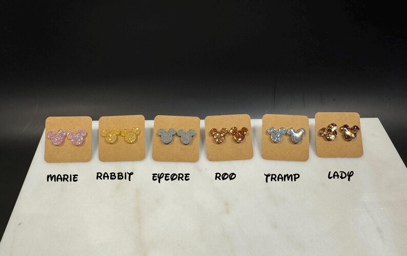 May include: Six pairs of glitter mouse ear stud earrings displayed on a white surface with labels for each pair: Marie, Rabbit, Eeyore, Roo, Tramp, Lady.