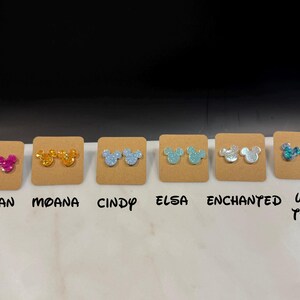 May include: Six pairs of glitter mouse ear stud earrings displayed on brown cards with the names Mulan, Moana, Cindy, Elsa, Enchanted, and Under the Sea printed below each pair.