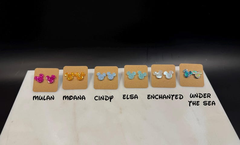 May include: Six pairs of glitter mouse ear stud earrings displayed on brown cards with the names Mulan, Moana, Cindy, Elsa, Enchanted, and Under the Sea printed below each pair.