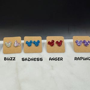 May include: Eight pairs of glitter Mickey Mouse earrings on brown cards. The cards are labeled with the names:  "Lotso", "Buzz", "Sadness", "Anger", "Rapunzel", and "Tangled".