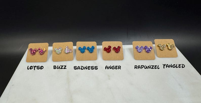 May include: Eight pairs of glitter Mickey Mouse earrings on brown cards. The cards are labeled with the names:  "Lotso", "Buzz", "Sadness", "Anger", "Rapunzel", and "Tangled".