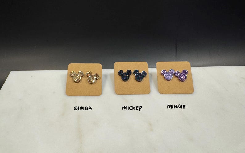 May include: Three pairs of glitter Mickey Mouse earrings on display cards. The earrings are gold, blue, and purple.
