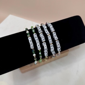 May include: Five beaded bracelets with white letter beads spelling out "FROG", "PASCAL", "SEE THE LIGHT", "GLEAM AND GLOW", and "BEST DAY EVER". The bracelets have green and clear beads and are displayed on a black velvet surface.
