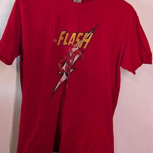 Könnte beinhalten: Rotes T-Shirt mit einer Grafik des Superhelden Flash. Das Design zeigt den Text "The Flash" in Gelb und eine rennende Figur auf einem Blitz. Das T-Shirt hängt an einem Holzkleiderbügel.