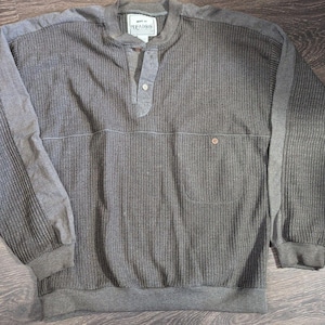 Peut inclure: Un pull Henley gris à manches longues avec une texture côtelée. Le pull est doté d'une patte de boutonnage à trois boutons, d'une poche poitrine et de panneaux gris contrastés sur les épaules et les manches. Les poignets et l'ourlet sont côtelés.