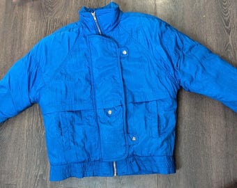May include: A bright blue jacket with a high collar and a zipper closure. The jacket has two front pockets and a buttoned flap. The sleeves have elastic cuffs. The jacket is made of a lightweight, possibly nylon, material.