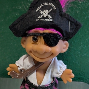 Puede incluir: Una muñeca troll vintage vestida de pirata, con un sombrero negro con el texto "Treasure Island at the Mirage", un parche en el ojo y una pipa marrón. La muñeca tiene el pelo rosa y lleva una camisa blanca y pantalones a cuadros.