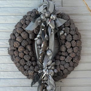 Peut inclure: Une couronne circulaire composée de nombreuses petites pommes de pin brunes et texturées. La couronne est ornée d'un arrangement vertical de rubans argentés, de coquillages peints en métal et de fines brindilles sombres torsadées.