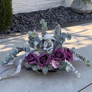 Puede incluir: Arreglo floral decorativo con rosas de color púrpura intenso, pequeñas flores rosas y detalles plateados. Una pequeña estatua de querubín se encuentra en el centro. El arreglo está sobre una superficie gris claro.