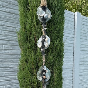 Op de afbeelding: Een verticale wanddecoratie bestaande uit vijf cirkelvormige donkergrijze vilten schijven op een rij. Elke schijf is versierd met een cluster van kleine witte en lichtgrijze schelpen. Het geheel hangt aan een dik, natuurlijk touw van jute.