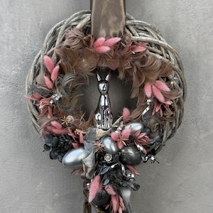 Könnte beinhalten: Ein dekorativer Osterkranz mit einem geflochtenen Weinreben-Sockel, verziert mit rosa und braunen Federn, silbernen Ostereiern und einer silbernen Hasenfigur. Ein braunes Satinband dient zum Aufhängen.