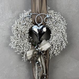 Könnte beinhalten: Ein dekorativer Kranz mit einem silbernen Herzanhänger und einem weißen Blumendesign. Der Kranz ist mit einem silbernen Drahtrahmen und einem braunen Stoffband verziert. Der Hintergrund ist einfarbig grau.