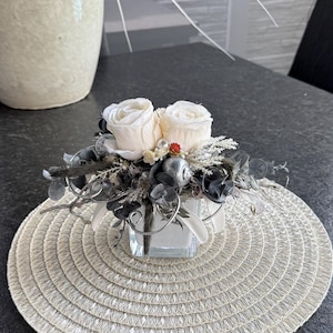 Könnte beinhalten: Ein kleines Blumenarrangement mit zwei weißen Rosen, dunkelgrauen und silbernen Akzenten, in einer quadratischen Glasvase, steht auf einem runden, beigefarbenen Platzset. Im Hintergrund steht ein großer, beigefarbener Keramiktopf mit einer Pflanze.