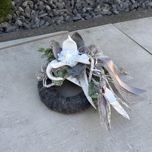 Op de afbeelding: Een grijze krans met een witte bloem en grijze lint accenten. De krans is versierd met gedroogde takken en groen.