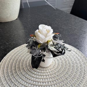 May include: A white rose floral arrangement in a small white pot, surrounded by dark gray and black accents. The arrangement sits on a woven, circular placemat. The placemat is on a dark gray surface, with a larger decorative pot in the background.