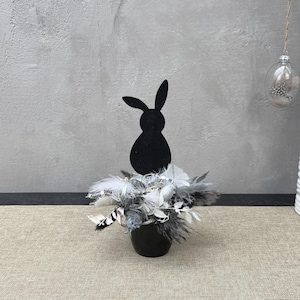 Può includere: Una silhouette nera di un coniglietto è posta su un piccolo vaso nero pieno di piume bianche e grigie. La composizione è su un runner da tavolo beige, con un muro grigio sullo sfondo. Un vaso bianco con rami e uova decorative è a destra.