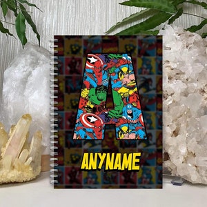 May include: A black spiral notebook with a colourful comic book pattern featuring superheroes. The letter "A" is formed by the comic book images. The text "ANYNAME" is printed in yellow at the bottom of the notebook.