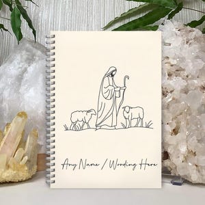 May include: A cream-colored spiral notebook with a black line drawing of a shepherd with two sheep. The text "Any Name / Wording Here" is at the bottom of the notebook.