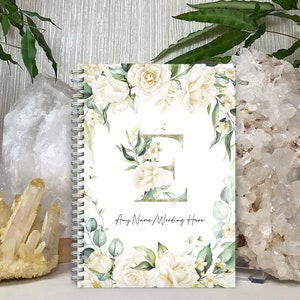 May include: White spiral bound notebook with a floral design featuring white roses and green leaves. The letter "E" is in the centre of the notebook with gold glitter. The text "Any Name/Wording Here" is at the bottom of the notebook.