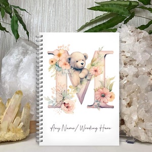 May include: A white spiral bound notebook featuring a pink and purple letter M adorned with watercolour flowers and a teddy bear. The text "Any Name/Wording Here" is at the bottom of the notebook.