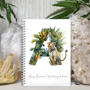 May include: A white spiral bound notebook featuring a watercolour illustration of a tiger in a jungle setting. The letter "A" is formed by the foliage. The text "Any Name / Wording Here" is printed at the bottom of the notebook.