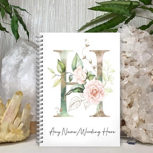 May include: A white spiral bound notebook featuring a gold and green watercolour letter H adorned with pink flowers. The text "Any Name/Wording Here" is printed at the bottom of the notebook.