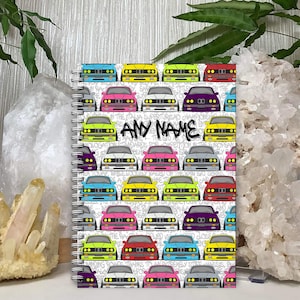 May include: A white spiral notebook with a colourful pattern of cartoon cars. The cars are in various colours, including red, yellow, blue, green, and pink. The text "ANY NAME" is printed on the notebook.