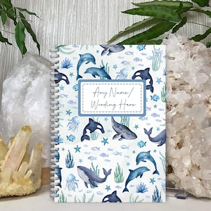 May include: A blue and white spiral bound notebook with a watercolour design of whales, dolphins, fish, and seaweed. The cover has a blue box with the text "Any Name/Wording Here".