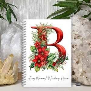 May include: A white spiral bound notebook featuring a red letter B surrounded by green leaves and red poinsettia flowers. The text "Any Name/Wording Here" is printed at the bottom of the notebook.