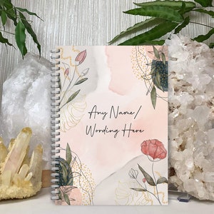 May include: A spiral-bound notebook with a watercolour design. The cover has a soft pink and grey background with gold, green, and red floral and botanical illustrations. Text on the cover reads "Any Name/ Wording Here".