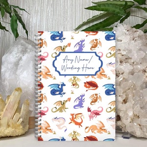 May include: A white spiral bound notebook with a colourful dragon pattern. The notebook has a blue label with the text "Any Name/Wording Here".