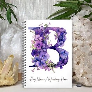 May include: A white spiral bound notebook featuring a purple watercolour letter B surrounded by purple flowers. The text "Any Name / Wording Here" is printed at the bottom of the notebook.