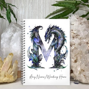 May include: A white spiral bound notebook featuring a watercolour illustration of a blue dragon forming the letter M. The text "Any Name/Wording Here" is printed at the bottom of the notebook.