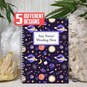 May include: A spiral bound notebook with a dark blue cover featuring a repeating pattern of planets, stars, comets, and moons in shades of yellow, orange, red, and purple. The text "Any Name/Wording Here" is printed on a purple rectangle in the centre of the cover.