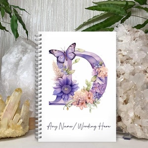 May include: A white spiral-bound notebook with a floral design. The cover displays a purple letter "D" decorated with flowers and a butterfly. The text "Any Name/Wording Here" is written at the bottom of the cover.