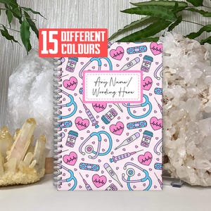 May include: A pink notebook with a spiral binding, decorated with a pattern of medical symbols in blue and white. The notebook has a label that says "Any Name/ Wording Here". A red rectangle above the notebook says "15 DIFFERENT COLOURS".