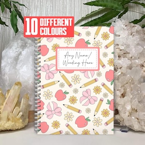 May include: A cream-coloured spiral-bound notebook with a repeating pattern of pink apples, bows, yellow pencils, and flowers. The notebook has a label with the text "Any Name/Wording Here". A red banner at the top reads "10 DIFFERENT COLOURS".