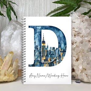 May include: A spiral-bound notebook with a white cover featuring a large letter D filled with a watercolour illustration of the New York City skyline, including the Statue of Liberty and skyscrapers. The text Any Name/Wording Here is printed below.